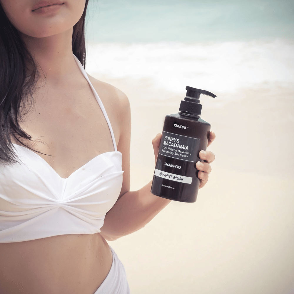 Donna in bikini bianco tiene KUNDAL Honey & Macadamia Nature Shampoo White Musk su una spiaggia con il mare sullo sfondo.