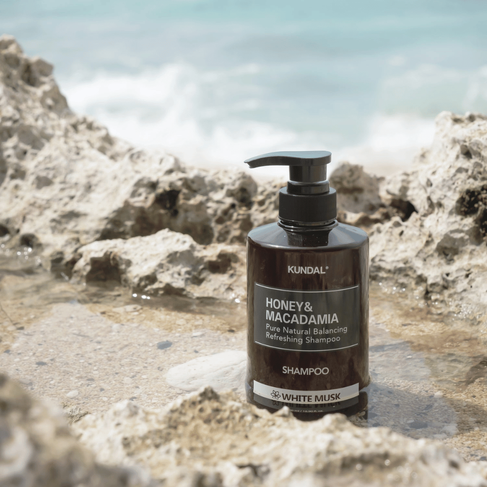KUNDAL Honey & Macadamia Nature Shampoo White Musk posizionata su scogli vicino al mare, un prodotto naturale per la cura dei capelli con un fresco profumo.
