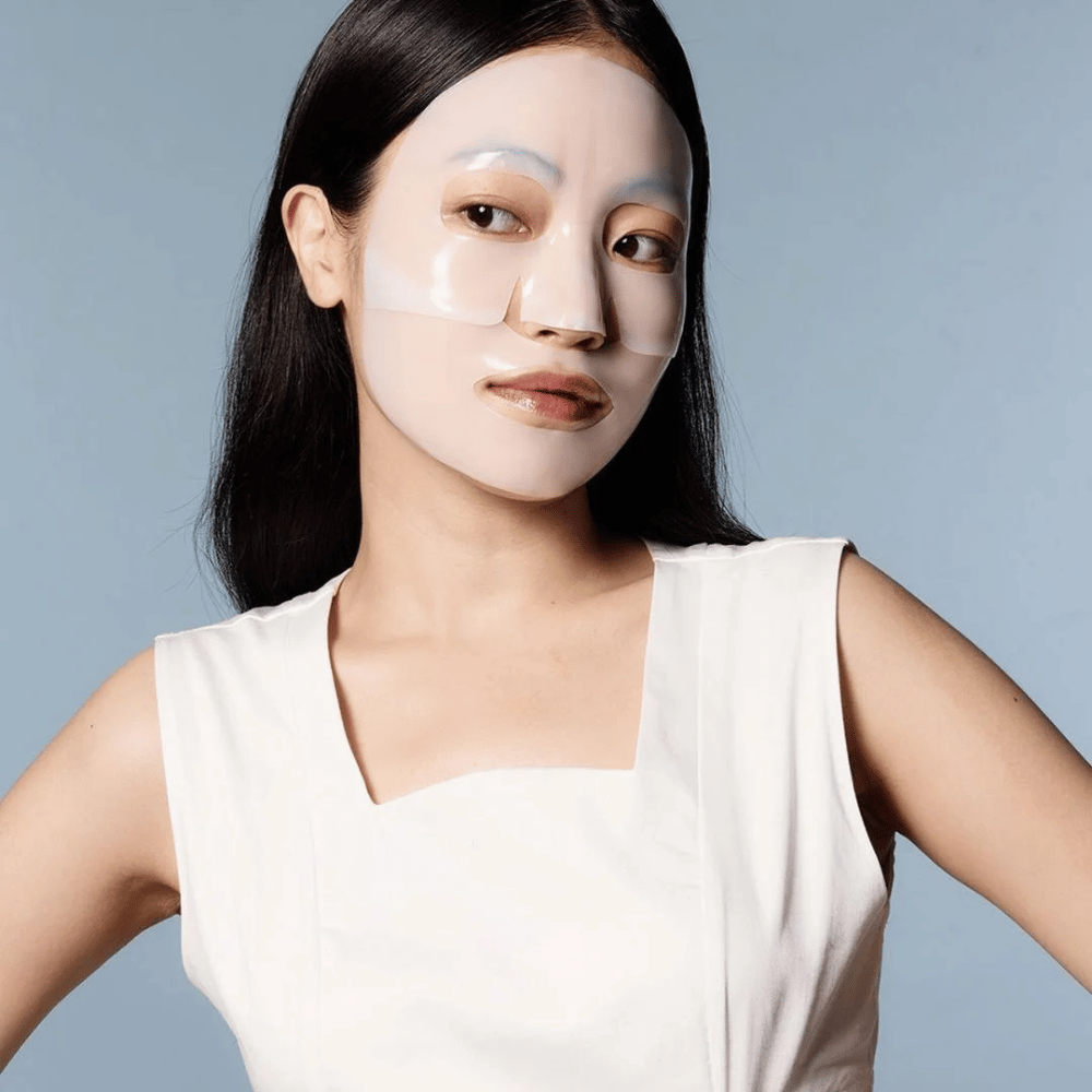 Una donna in un abito bianco senza maniche indossa un maschera viso trasparente che copre quasi tutto il suo viso. La maschera dà un'impressione di cura della pelle avanzata e offre l'aspetto di un trattamento di bellezza, progettata per idratare in profondità e trattare la pelle.
