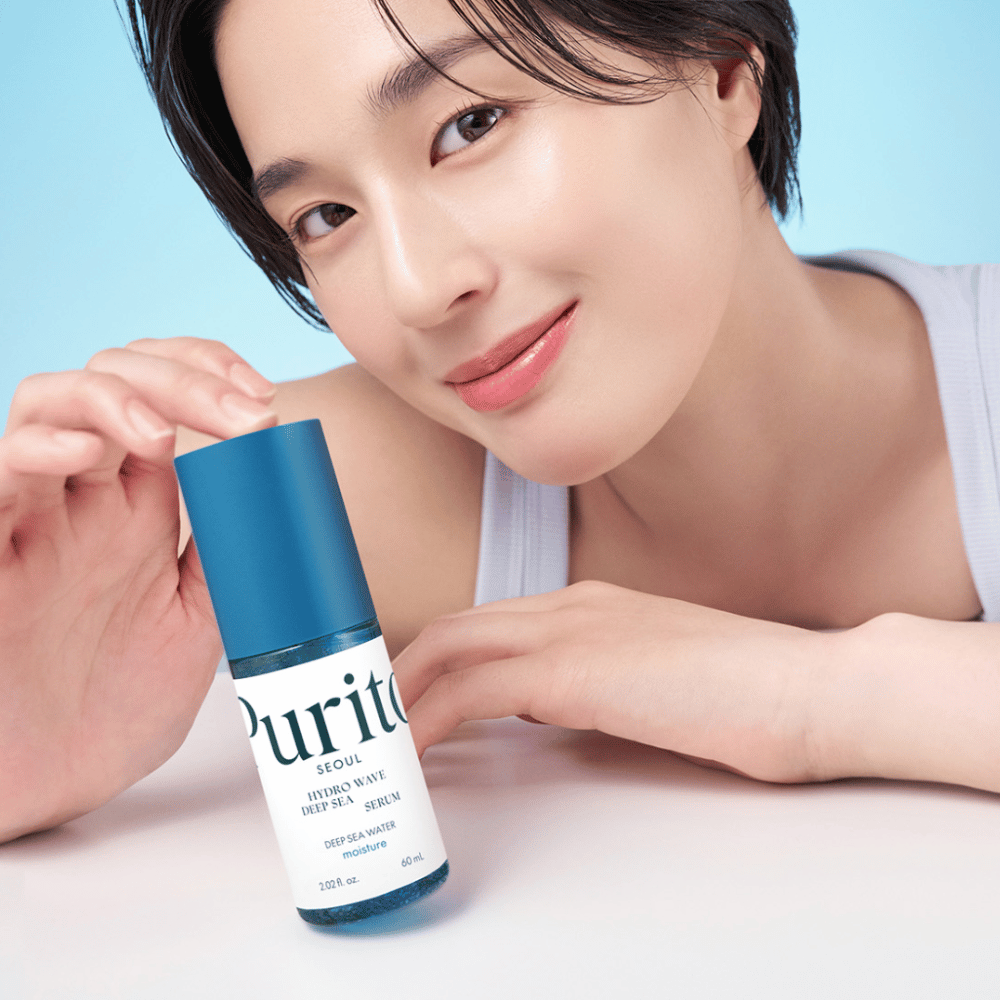 Una donna con capelli corti tiene un siero blu di Purito, Hydro Wave Deep Sea Serum. La sua pelle appare radiosa e idratata, mettendo in risalto il prodotto in primo piano. Lo sfondo è azzurro chiaro, che dona una sensazione fresca e pulita. Il prodotto è focalizzato sulla cura della pelle.