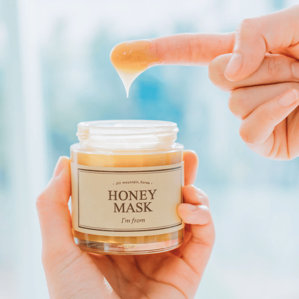 Una mano tiene un barattolo aperto di "I'm from Honey Mask", e un dito viene immerso nella maschera dorata su uno sfondo chiaro.