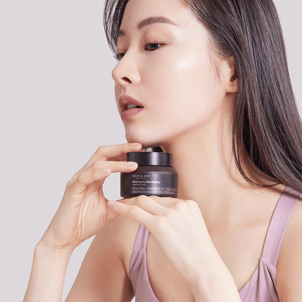 Una donna tiene in mano un barattolo di MARY & MAY Idebenone Blackberry Intense Cream. Ha una pelle radiosa e sorride leggermente, vestita con un top senza maniche. Il prodotto è una crema intensa con idebenone e complesso di more, destinata alla compattezza e idratazione della pelle.