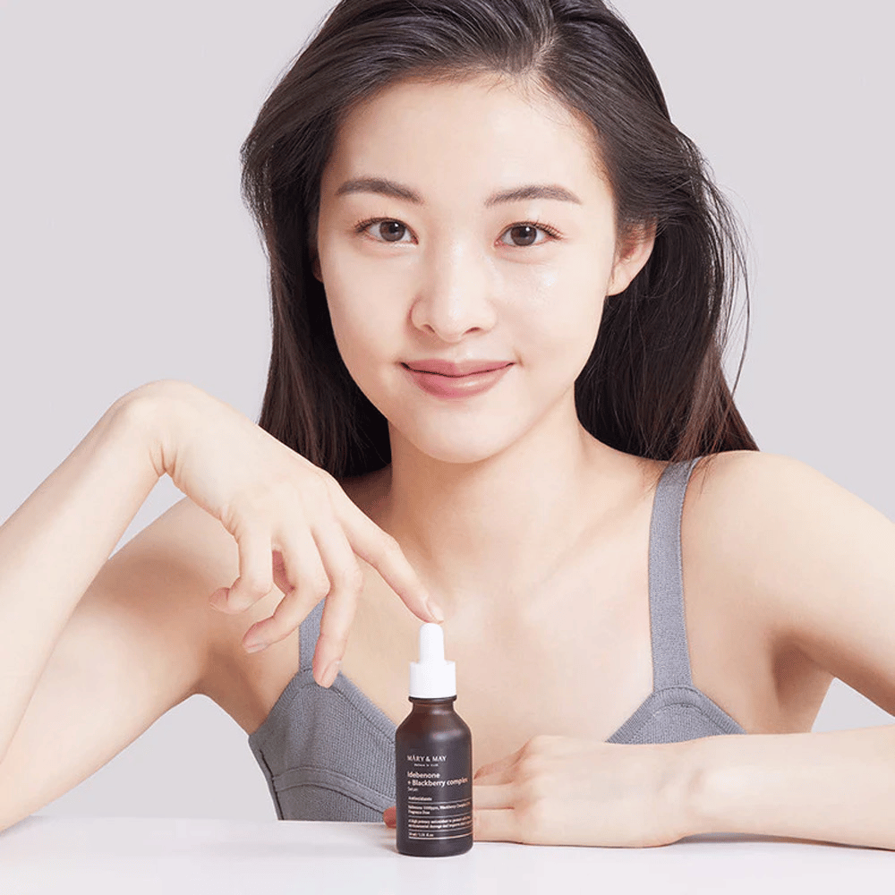 Una donna con lunghi capelli scuri tiene in mano MARY & MAY Idebenone + Blackberry Complex Serum, una bottiglia di vetro scuro con pipetta. Ha un sorriso felice e indica la bottiglia con l'altra mano. La donna è vestita con un top grigio senza maniche e sembra trovarsi in un ambiente luminoso e semplice, il che mette in risalto l'attenzione del prodotto per la cura della pelle e la bellezza.