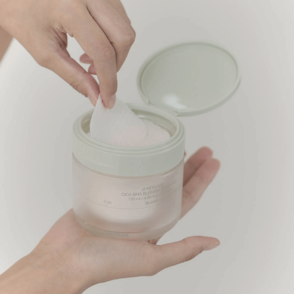 Una mano prende un pad tonico viso dal barattolo Celimax Ji Woo Gae Cica BHA Blemish Toner Pad, perfetto per la pelle impura.