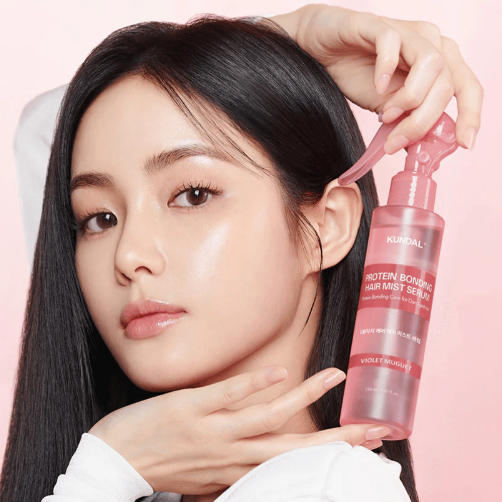 Donna con lunghi capelli neri tiene in mano KUNDAL Protein Bonding Hair Mist Serum Violet Muguet davanti al viso, su uno sfondo rosa chiaro.