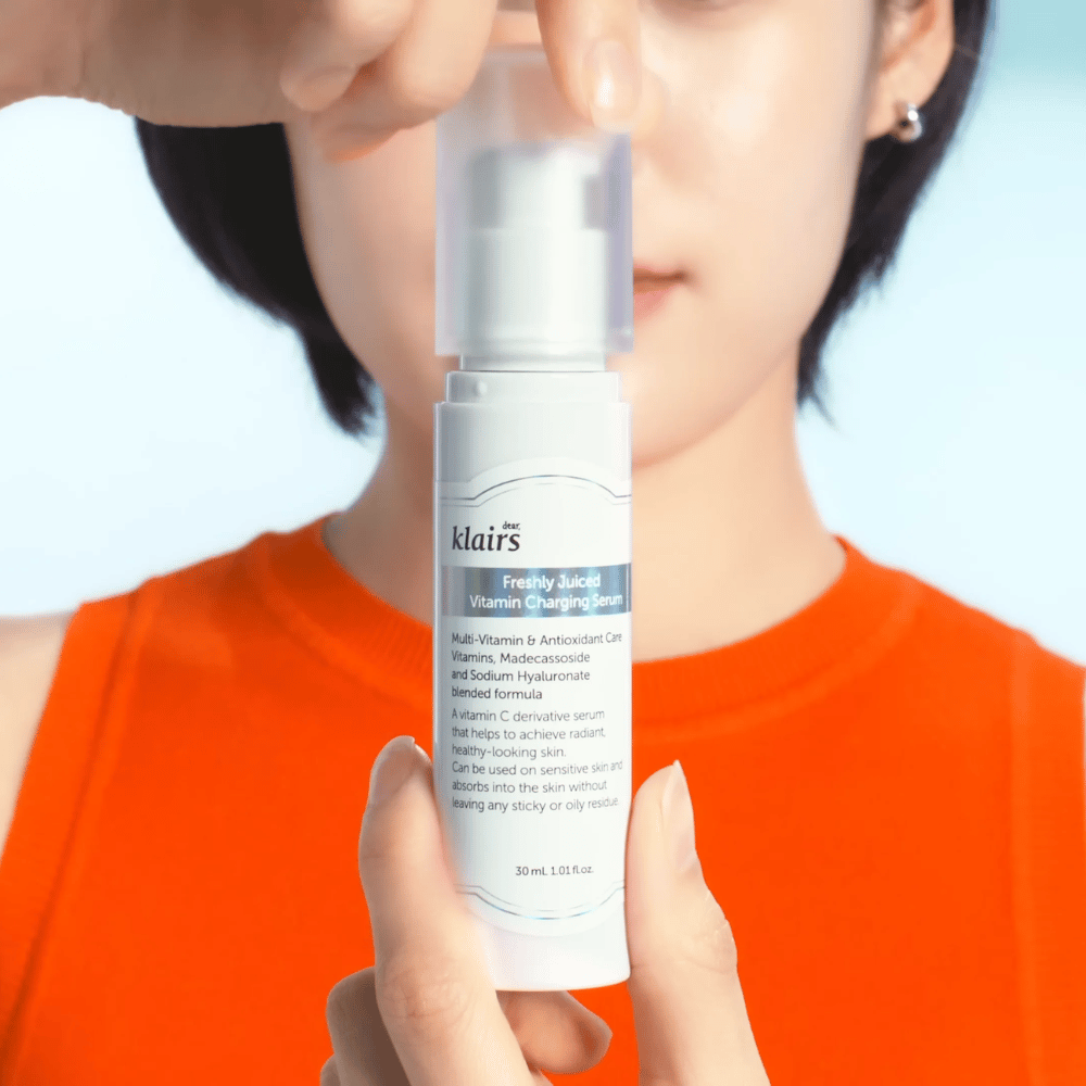 Donna in maglia arancione tiene Klairs Freshly Juiced Vitamin Charging Serum davanti a sé su uno sfondo blu.