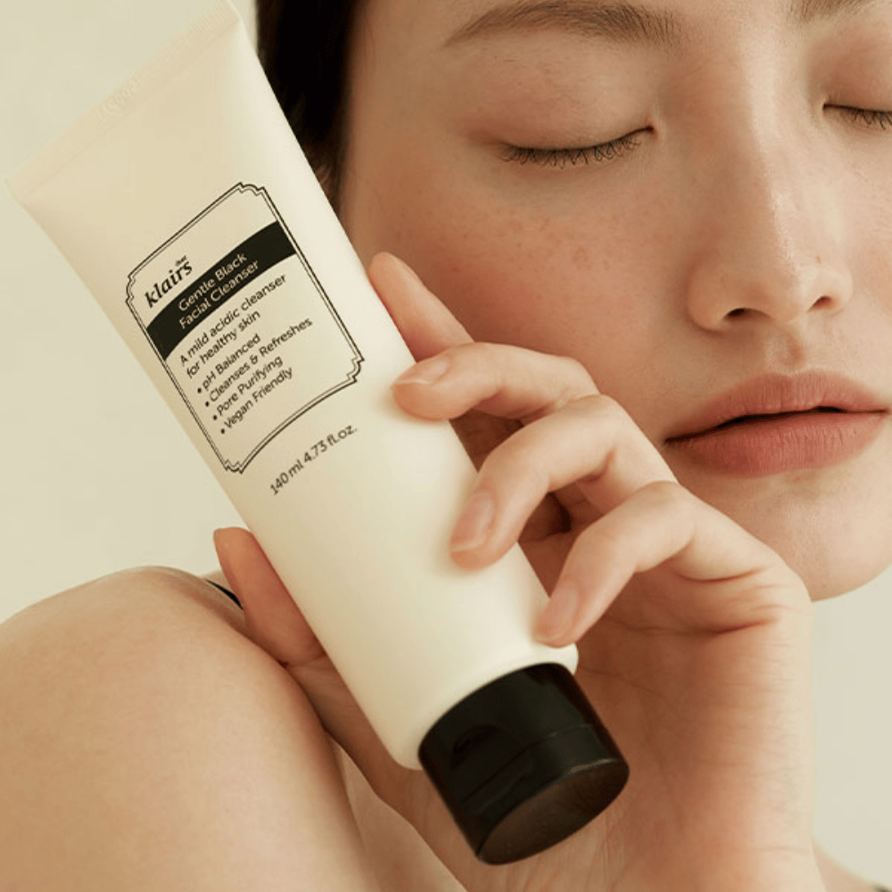Una persona tiene un tubo di Klairs Gentle Black Facial Cleanser contro la sua guancia, occhi chiusi e tono della pelle naturale visibili sullo sfondo.