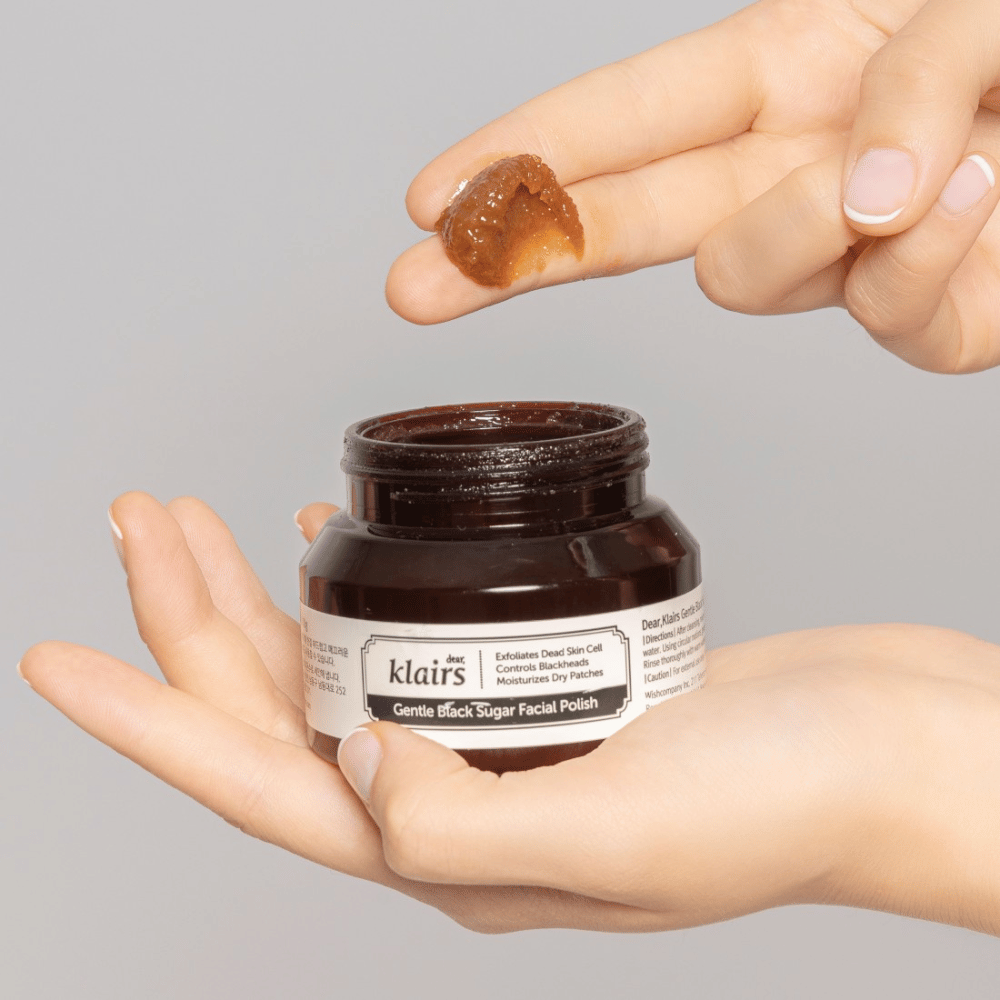 Mani tengono un barattolo aperto di Klairs Gentle Black Sugar Facial Polish e mostrano il prodotto su un dito, su uno sfondo grigio.