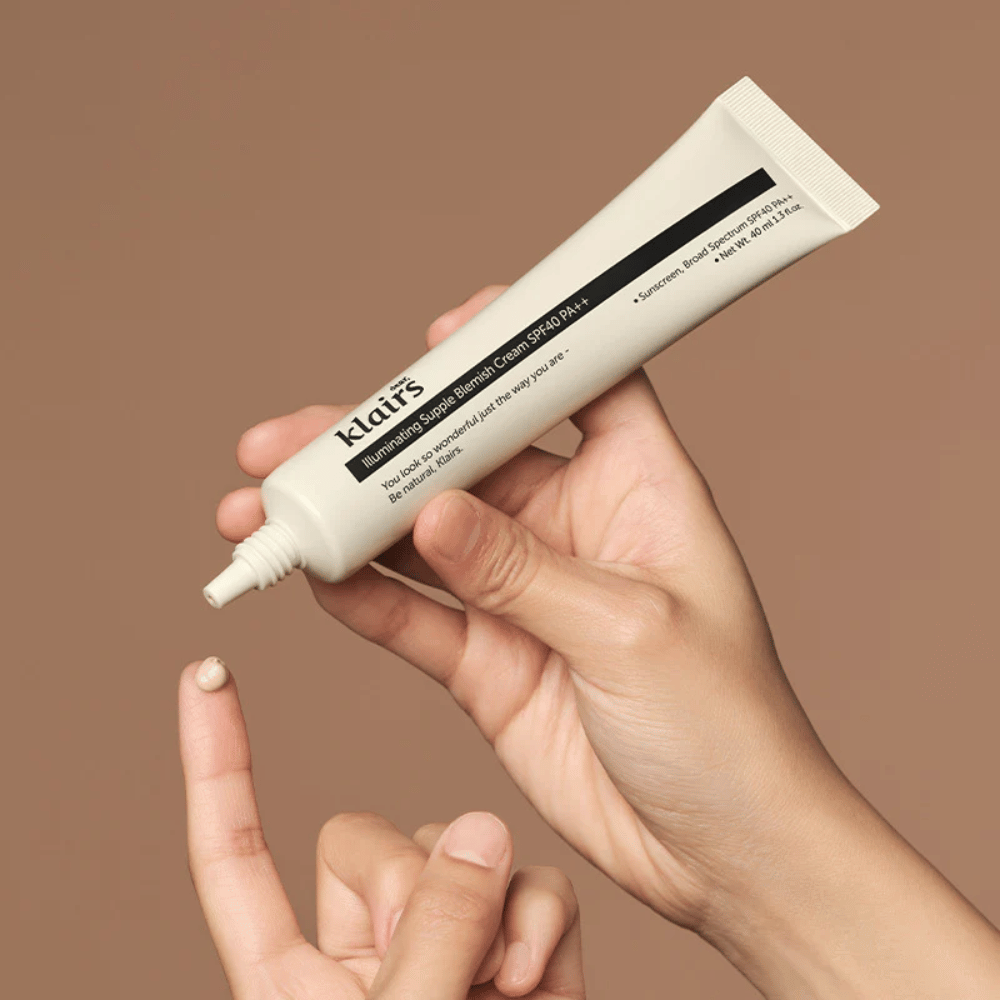 Una mano tiene un tubo di Klairs Illuminating Supple Blemish Cream SPF40 PA++ e preme una piccola quantità di crema su un dito, su uno sfondo marrone.