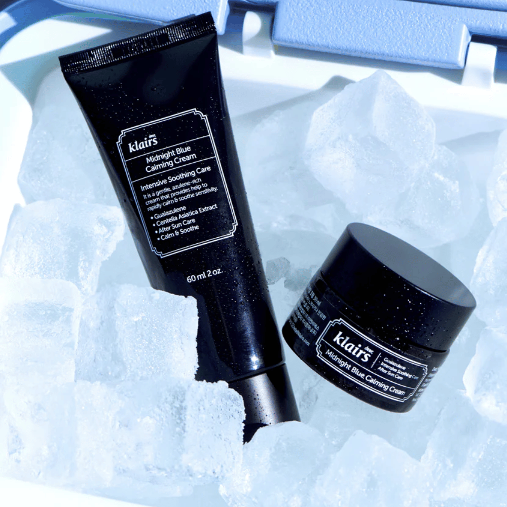 Un tubo e un barattolo di Klairs Midnight Blue Calming Cream si trovano su cubetti di ghiaccio, con un focus su proprietà lenitive e idratanti, contro uno sfondo blu.