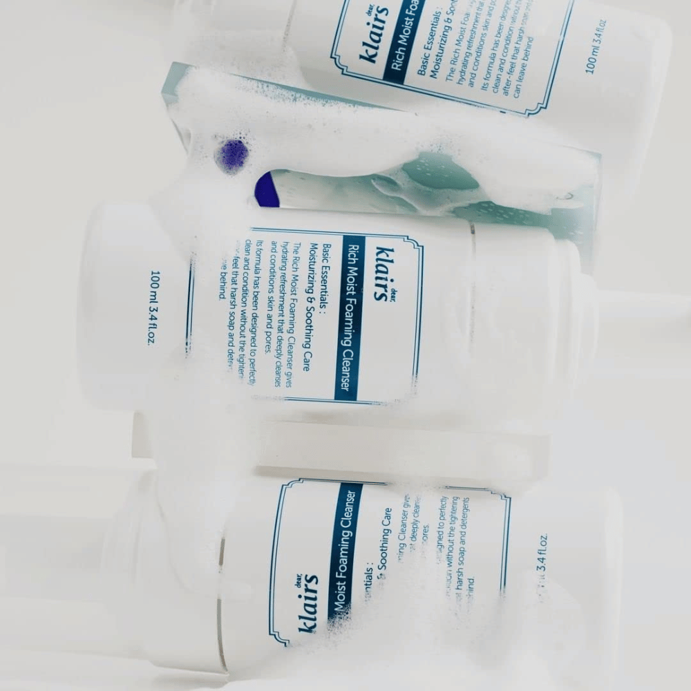 Tre flaconi di Klairs Rich Moist Foaming Cleanser coperti di schiuma e gocce d'acqua, posizionati su una superficie bianca.