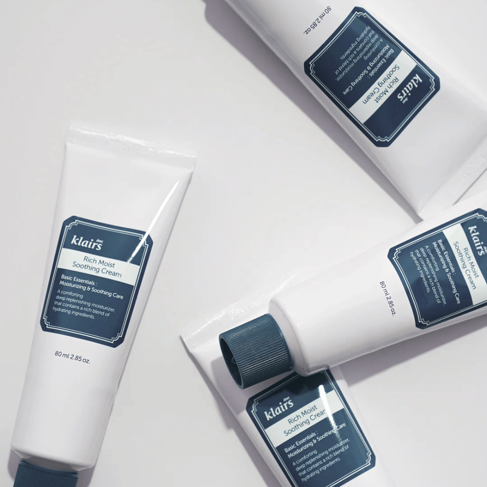 Diversi tubi di Klairs Rich Moist Soothing Cream posizionati strategicamente su una superficie chiara, con ombre che creano profondità.