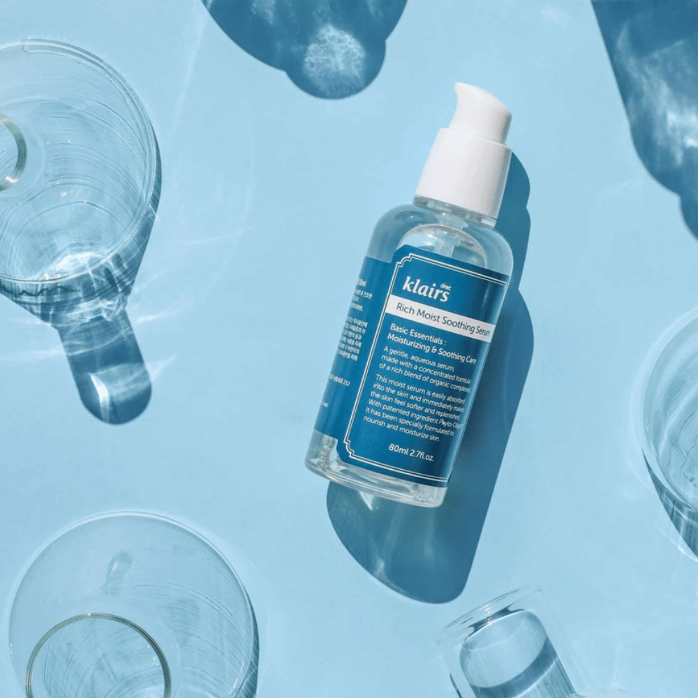 Una bottiglia Klairs Rich Moist Soothing Serum tra bicchieri d'acqua con riflessi su uno sfondo blu.