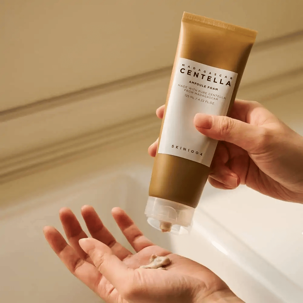 Il prodotto nell'immagine è "Madagascar Centella Ampoule Foam" di SKIN1004. È un detergente viso sotto forma di schiuma, realizzato con Centella del Madagascar. Contiene 125 ml / 4.22 fl.oz. Una mano preme una piccola quantità di prodotto per l'uso.