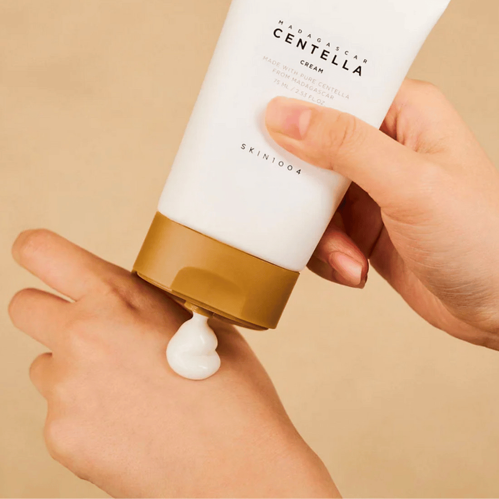 L'immagine mostra una persona che tiene in mano un tubo di "Madagascar Centella Cream" da SKIN1004. È una crema per la pelle realizzata con pura Centella del Madagascar, nella dimensione di 75 ml / 2.53 fl.oz. La crema viene applicata sulla mano ed è destinata a fornire nutrimento e idratazione alla pelle.