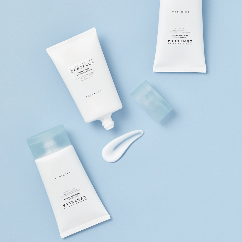 Due tubi bianchi con SKIN1004 prodotti per la cura della pelle Centella, una noce di prodotto e un tappo blu trasparente, su uno sfondo azzurro.