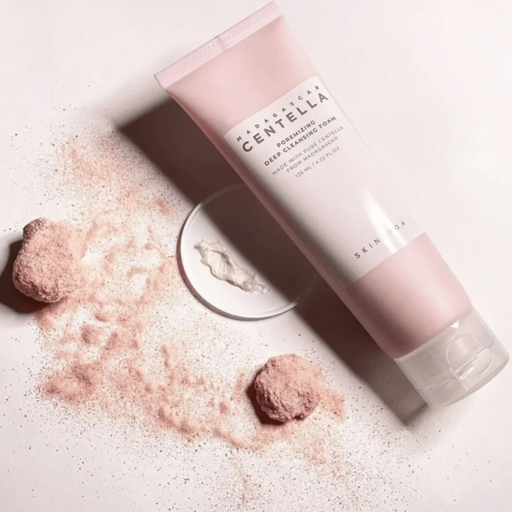 Madagascar Centella Poremizing Deep Cleansing Foam è presentato in un tubetto rosa. I tubetti sono posizionati su una superficie chiara insieme a polvere rosa che conferisce una sensazione di pulizia e freschezza. Il prodotto è progettato per pulire in profondità la pelle con l'aiuto di centella pura del Madagascar.
