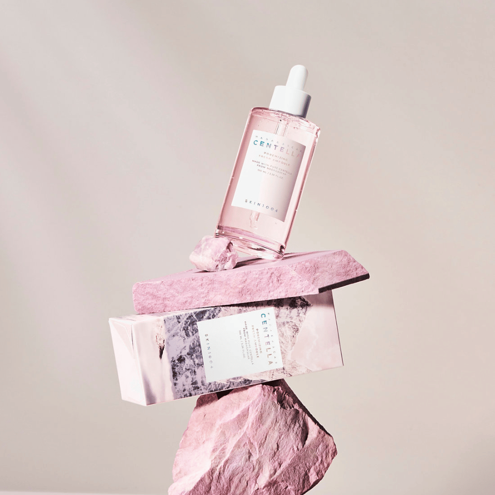 Bottiglia trasparente con ampolla rosa Centella da SKIN1004, bilanciata su blocchi di pietra, con una confezione di cartone sotto, su uno sfondo neutro.