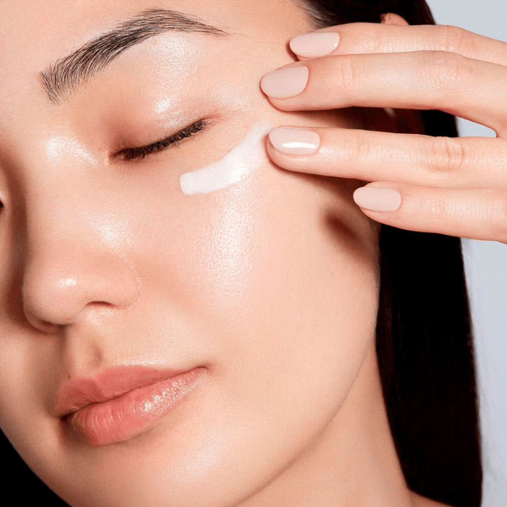 Una donna applica delicatamente una crema contorno occhi bianca sotto l'occhio. La sua pelle appare radiosa e idratata, il che sottolinea l'effetto della crema per gli occhi. Questo mette in risalto il prodotto come sia idratante che adatto per la pelle sensibile intorno agli occhi.