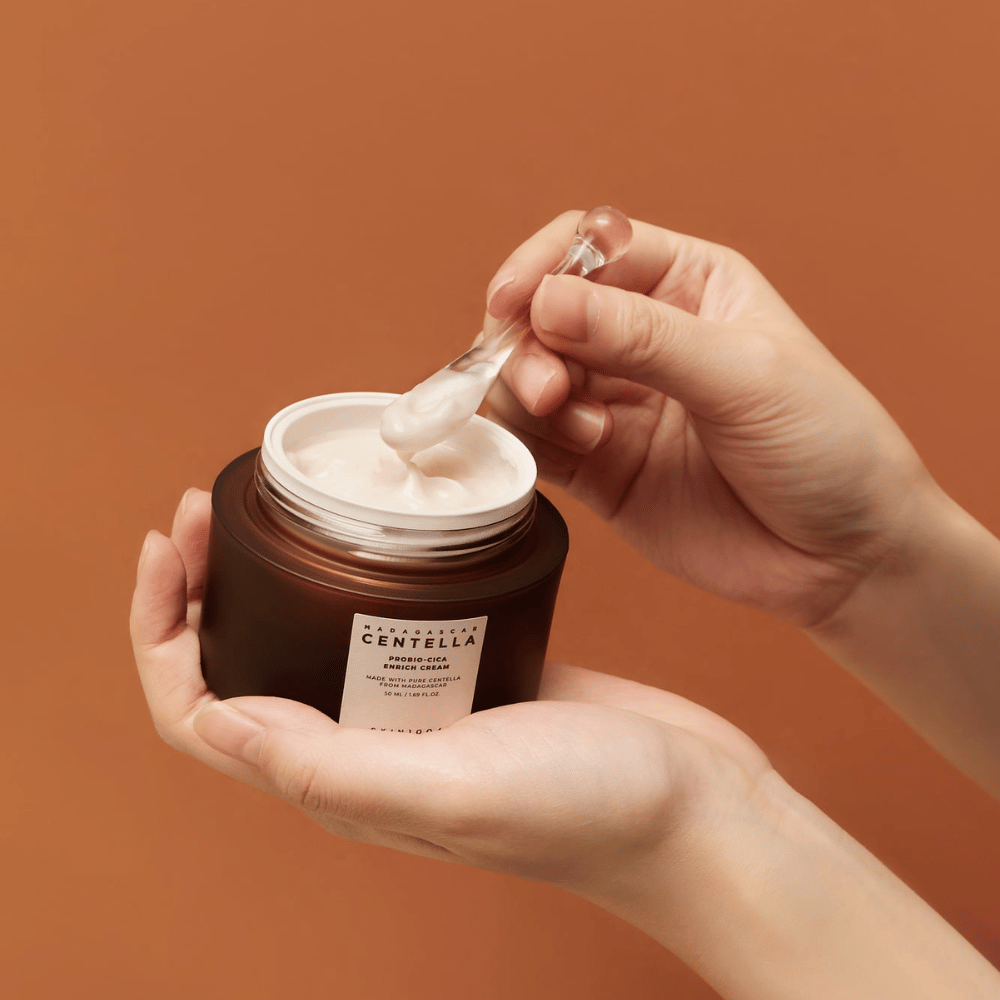 Mani che tengono un barattolo aperto di Madagascar Centella Probio-Cica Enrich Cream e una spatola con crema su uno sfondo uniforme color terracotta.