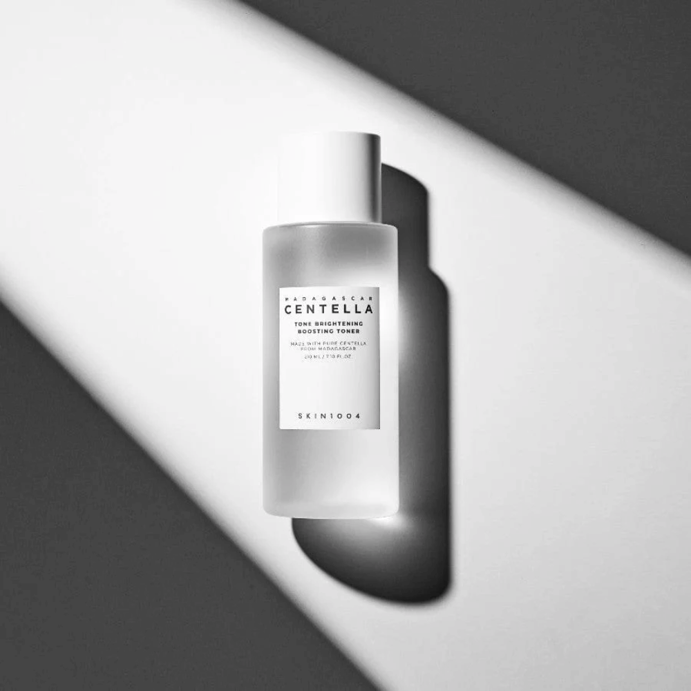 Bottiglia di SKIN1004 Madagascar Centella Tone Brightening Boosting Toner  sdraiata su una superficie bianca con un'ombra drammatica in gioco di luci e ombre in bianco e nero.