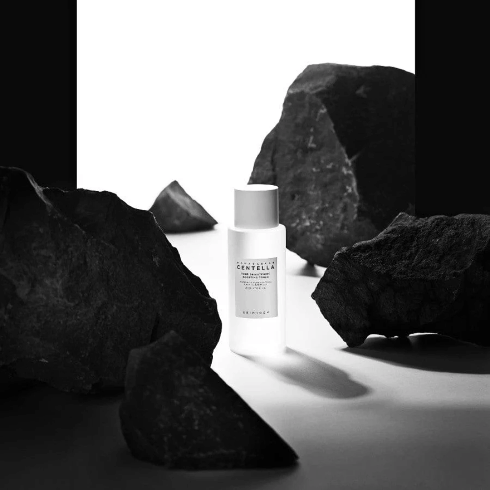 Bottiglia bianca SKIN1004 Madagascar Centella Tone Brightening Boosting Toner  posizionata drammaticamente tra grandi pietre nere su sfondo bianco.