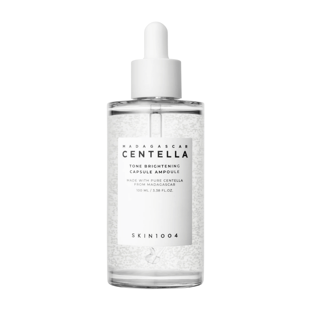L'immagine mostra una bottiglia di "SKIN1004 Madagascar Centella Tone Brightening Capsule Ampoule". È una bottiglia trasparente con un applicatore a goccia bianco. Sull'etichetta si legge che il prodotto contiene ingredienti puri di centella dal Madagascar. Il contenuto della bottiglia sembra avere piccole capsule o particelle, che potrebbero far parte delle sue proprietà illuminanti per la cura della pelle. La bottiglia contiene 100 ml / 3.38 fl.oz. di prodotto.