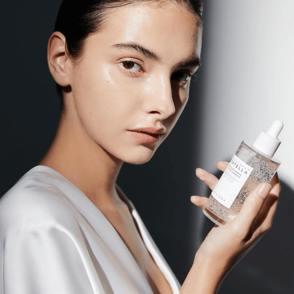 Nell'immagine si vede una persona che tiene una bottiglia con applicatore a goccia. La bottiglia è etichettata "SKIN1004 Madagascar Centella Tone Brightening Capsule Ampoule". La persona ha un tono di pelle scintillante che riflette la luce, suggerendo un possibile effetto del prodotto.