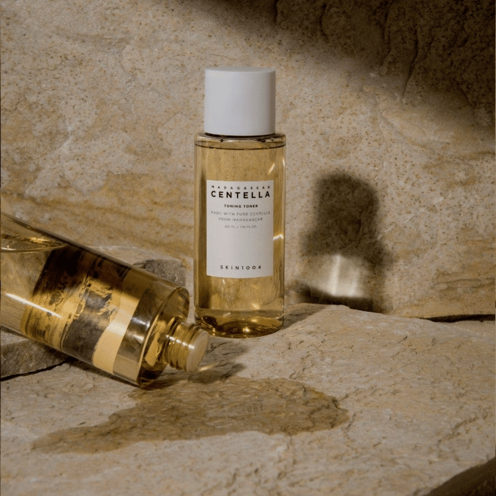 L'immagine mostra una bottiglia "Madagascar Centella Toning Toner" da SKIN1004. È una bottiglia trasparente riempita con un liquido giallastro, probabilmente un tonico viso per la cura della pelle. Si dice che sia fatta con pura centella del Madagascar e il contenuto è di 210 ml / 7.10 fl.oz. La bottiglia e una bottiglia sdraiata sono posizionate su una superficie rocciosa, che conferisce una sensazione naturale e minimalista.