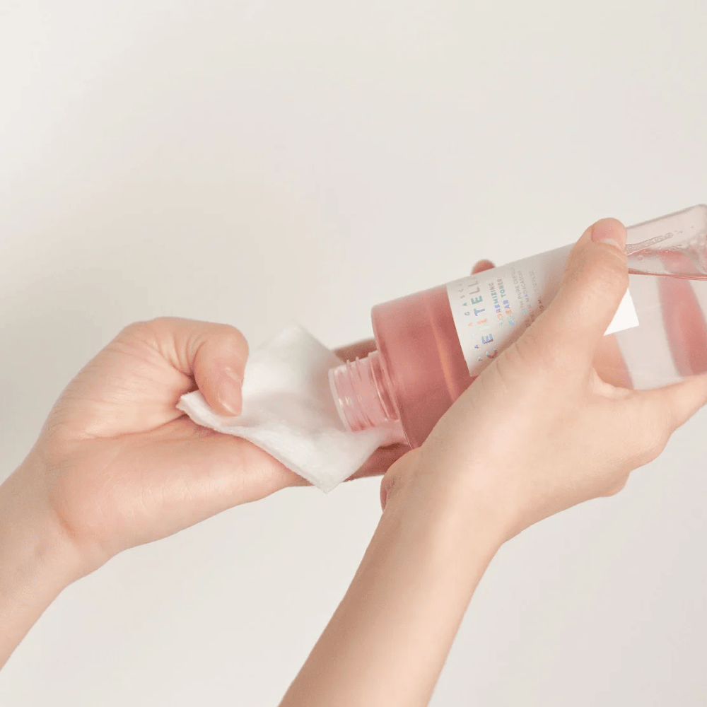 Una persona versa Madagascar Centella Poremizing Clear Toner da una bottiglia trasparente su un dischetto di cotone. Il prodotto, che ha una tonalità rosa, viene utilizzato per rinfrescare e pulire la pelle. Le mani sono visibili, mostrando l'applicazione del toner.