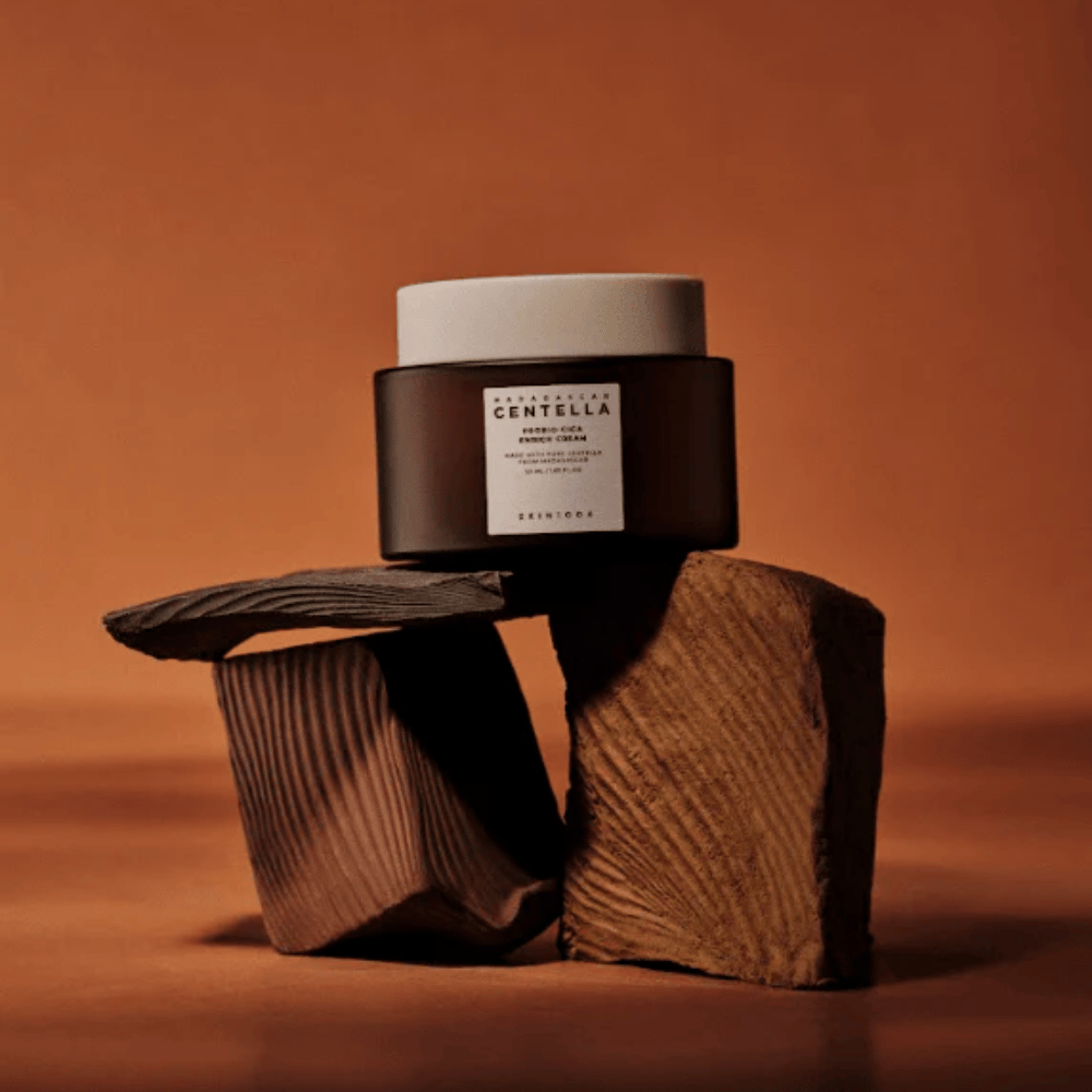 SKIN1004 Madagascar Centella Probio-Cica Enrich Cream in elegante barattolo di vetro marrone su blocchi di legno decorativi – crema viso coreana nutriente che rafforza la barriera cutanea.