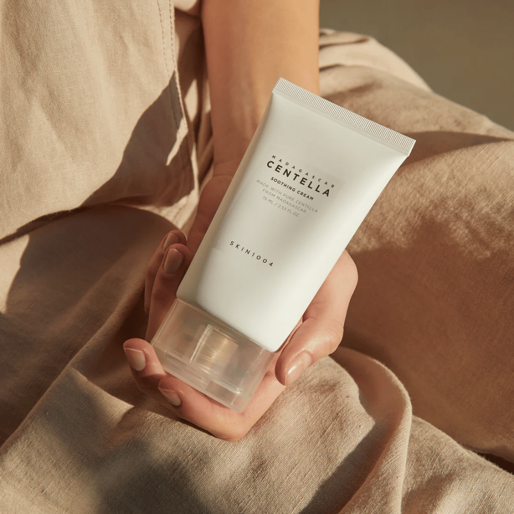 Una persona tiene un tubo bianco di Madagascar Centella Soothing Cream da SKIN1004. Il tubo ha un tappo trasparente e un testo che descrive il prodotto come calmante e realizzato con pura Centella. La persona è vestita con abiti chiari.