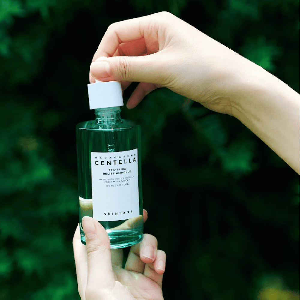 Persona apre SKIN1004 Madagascar Centella Tea-Trica Relief Ampoule – bottiglia di vetro verde con etichetta bianca tenuta in mano davanti a uno sfondo rigoglioso.