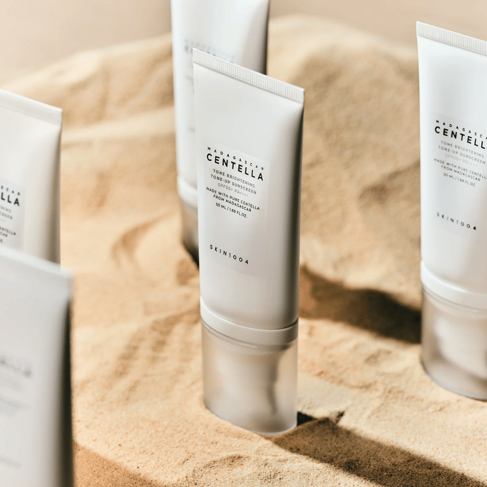 Diversi tubetti di Madagascar Centella Tone Brightening Tone-Up Sunscreen sono nella sabbia. Il tubetto è bianco con testo nero e contiene 50 ml. Il prodotto ha SPF 50+ PA++++ ed è realizzato con pura Centella del Madagascar.