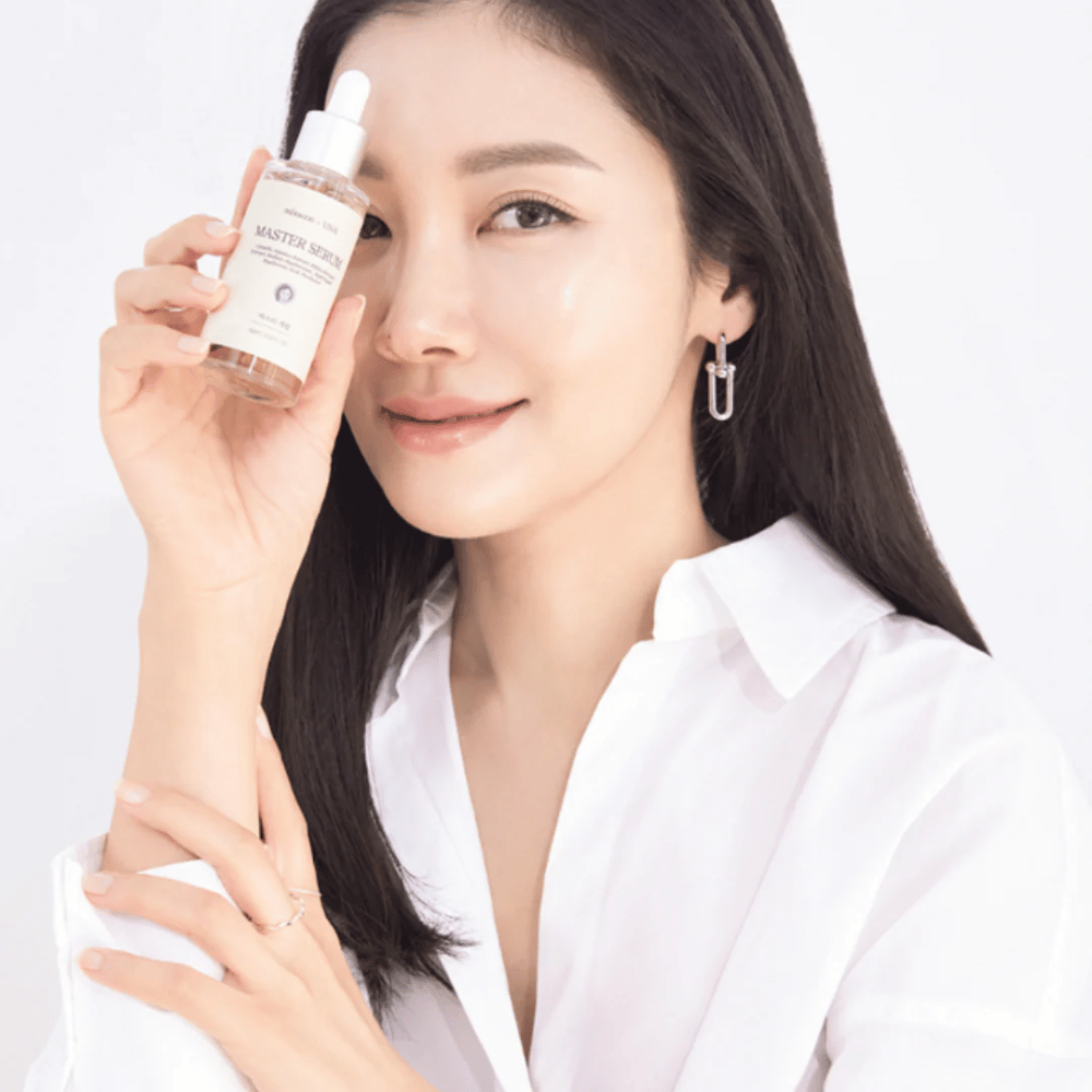 Donna sorridente con capelli lunghi e scuri e camicia bianca tiene Mixsoon Master Serum accanto al suo viso. La sua pelle appare radiosa e idratata.