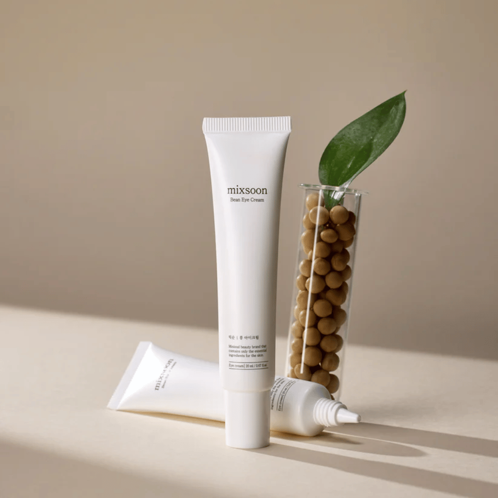 Due tubi Mixsoon Bean Eye Cream posizionati accanto a una provetta riempita di semi di soia e una foglia verde, su una superficie beige con giochi di ombre.