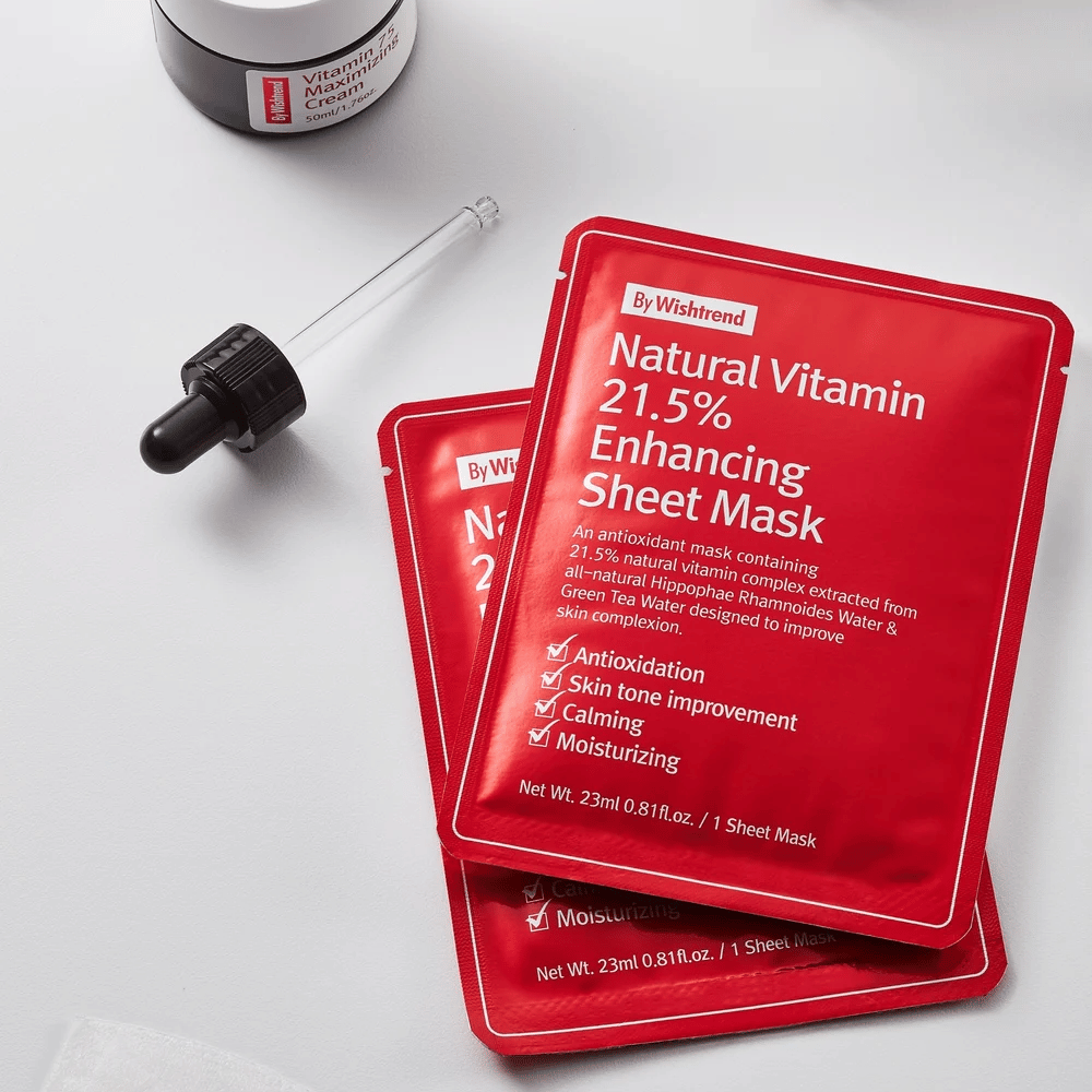 By Wishtrend Natural Vitamin 21.5% Enhancing Sheet Mask in confezioni rosse, posizionate su una superficie bianca insieme a una pipetta e un barattolo di crema per il viso.