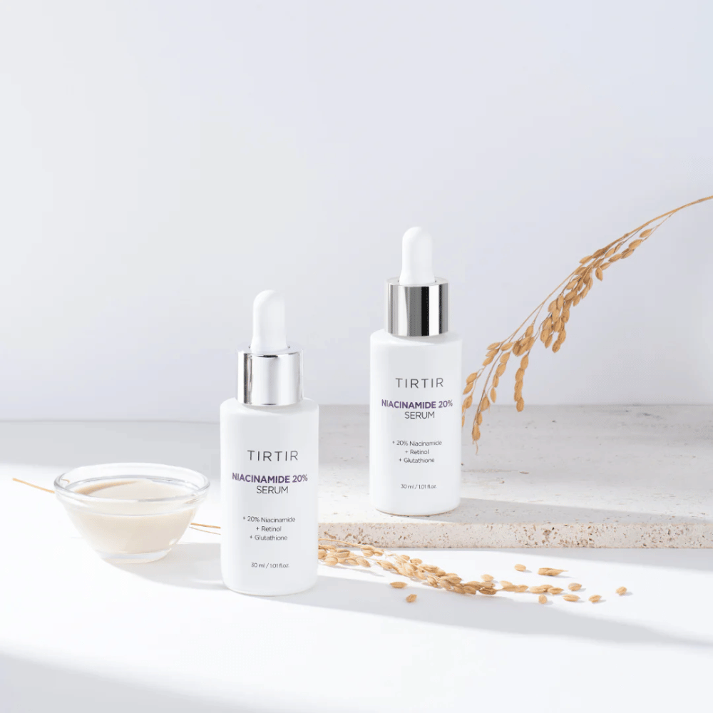 Due bottiglie di Tirtirs "Niacinamide 20% Serum" sono mostrate su una superficie chiara insieme a una piccola ciotola con una sostanza cremosa e alcuni fili di piante secche. Il siero contiene il 20% di niacinamide, retinolo e glutatione, il che aiuta a migliorare l'aspetto e la texture della pelle.