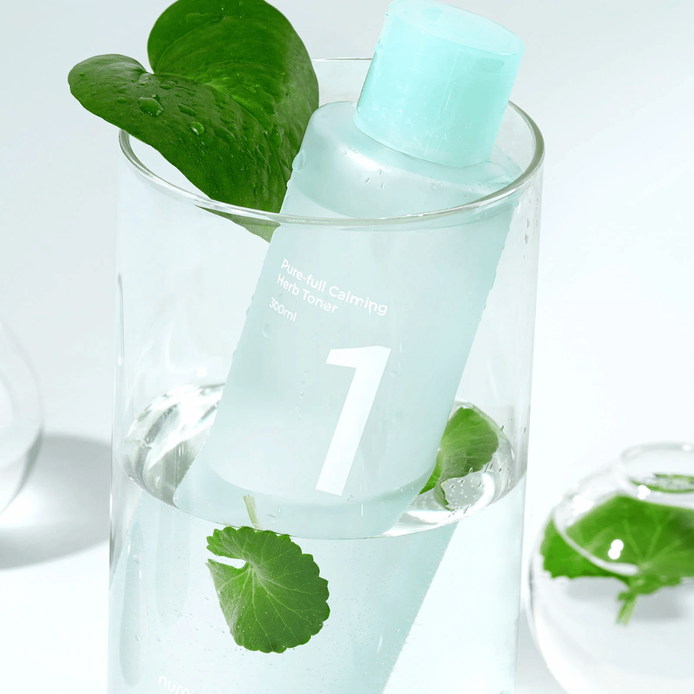 Una bottiglia con No.1 Pure-Full Calming Herb Toner posizionata in un bicchiere con acqua e foglie verdi. La bottiglia ha un tappo azzurro chiaro e un'etichetta con il testo "Pure-full Calming Herb Tonico viso" e "300ml". Il prodotto appare fresco con acqua e vegetazione sullo sfondo, il che conferisce una sensazione rilassante e naturale.