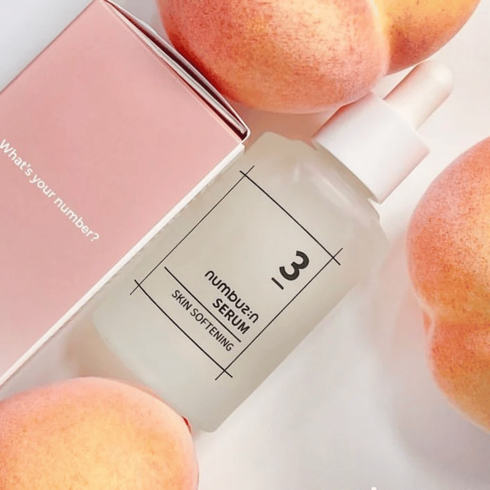 Bottiglia con numbus:in No.3 Skin Softening Serum circondata da pesche e una scatola rosa con il testo "What's your number?".