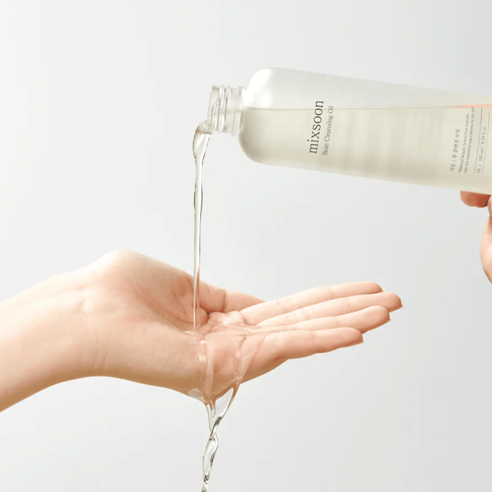 Un olio detergente chiaro viene versato da una bottiglia trasparente con etichetta bianca dove è scritto "MixSoon" in un palmo aperto. L'olio forma un flusso lucente tra l'apertura della bottiglia e la mano, dimostrando la consistenza liquida dell'olio. La mano è ferma e viene tenuta in modo da raccogliere l'intera quantità di olio detergente contro uno sfondo chiaro e uniforme.