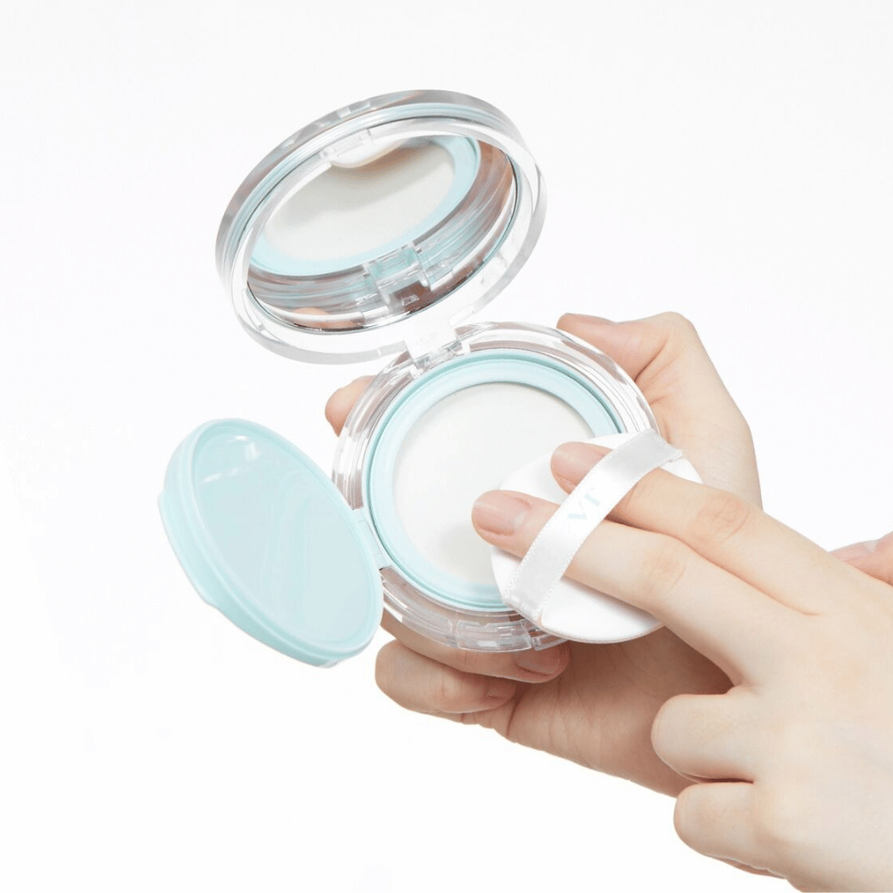 Una mano tiene un barattolo aperto con VT PDRN Essence Glow Sun Pact. Il puff sta per applicare la polvere, che ha una superficie bianca e un bordo blu attorno al barattolo.