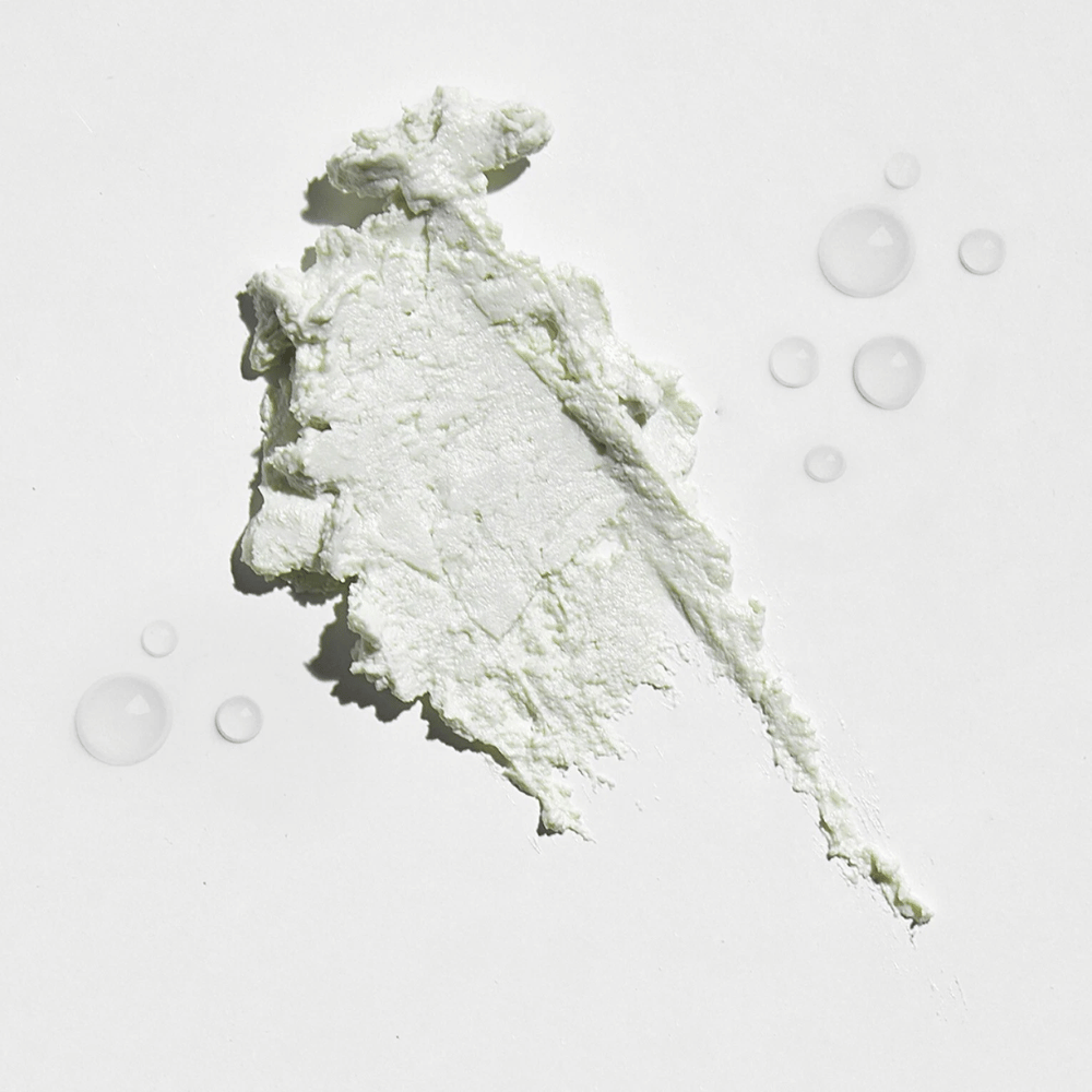 Primo piano di una sostanza bianca e cremosa su uno sfondo bianco, con alcune gocce d'acqua intorno. La texture sembra essere una terra abbronzante o un fondotinta con una leggera luminosità.
