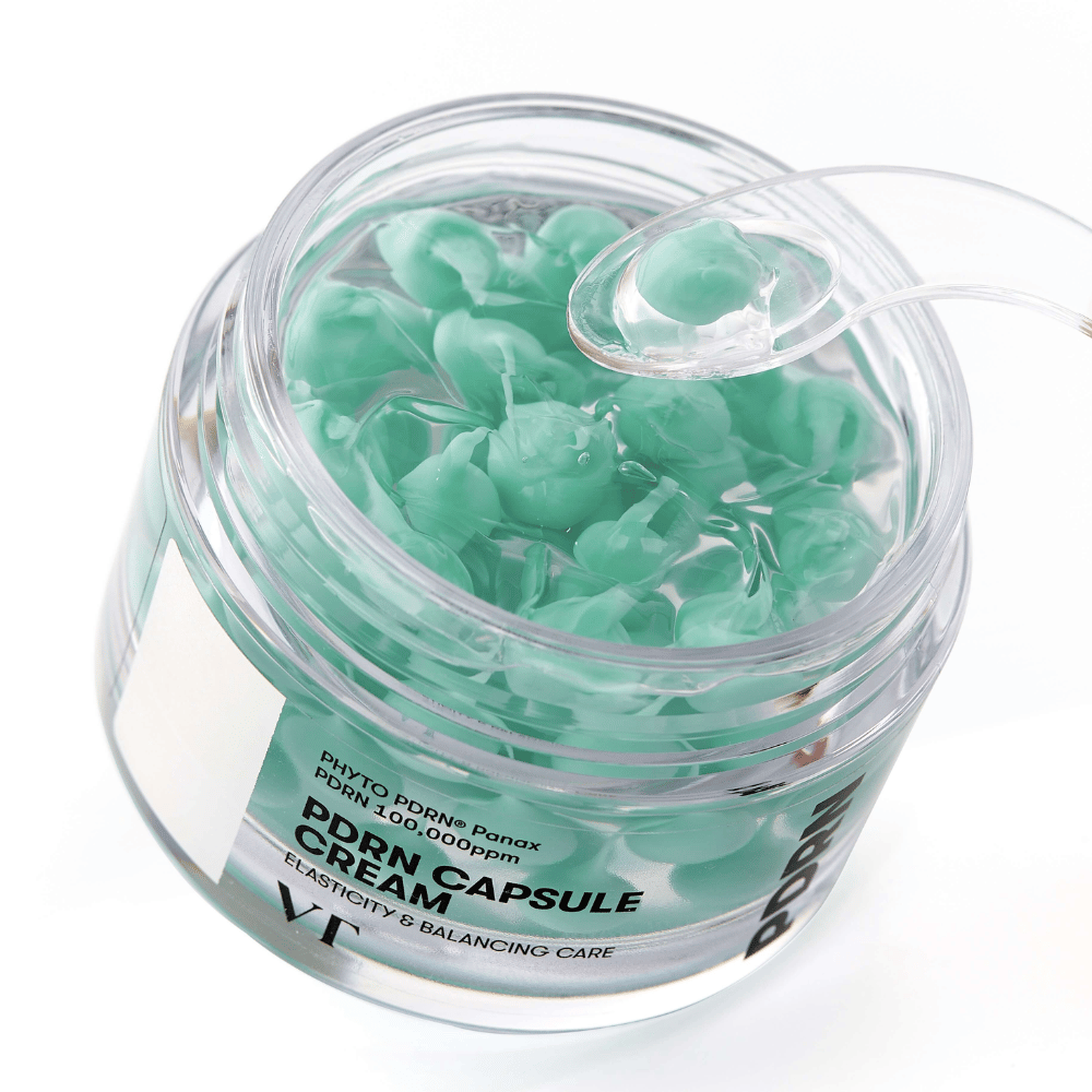 Primo piano di un barattolo aperto con VT PDRN Capsule Cream. Il contenuto è composto da piccole capsule verdi in un gel trasparente, con una spatola inclusa per l'applicazione. Il testo sul barattolo recita "Elasticity & Balancing Care".