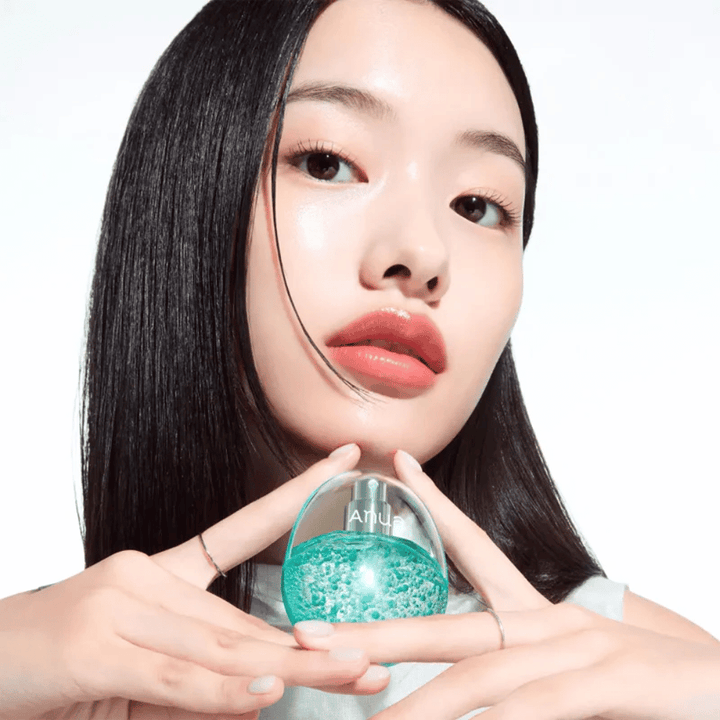 Primo piano di una modella che tiene una bottiglia rotonda e trasparente di Anua riempita con capsule o perle di gel color turchese. L’immagine si concentra sulla luminosità della pelle e sul design innovativo del prodotto, trasmettendo un’impressione fresca e moderna.