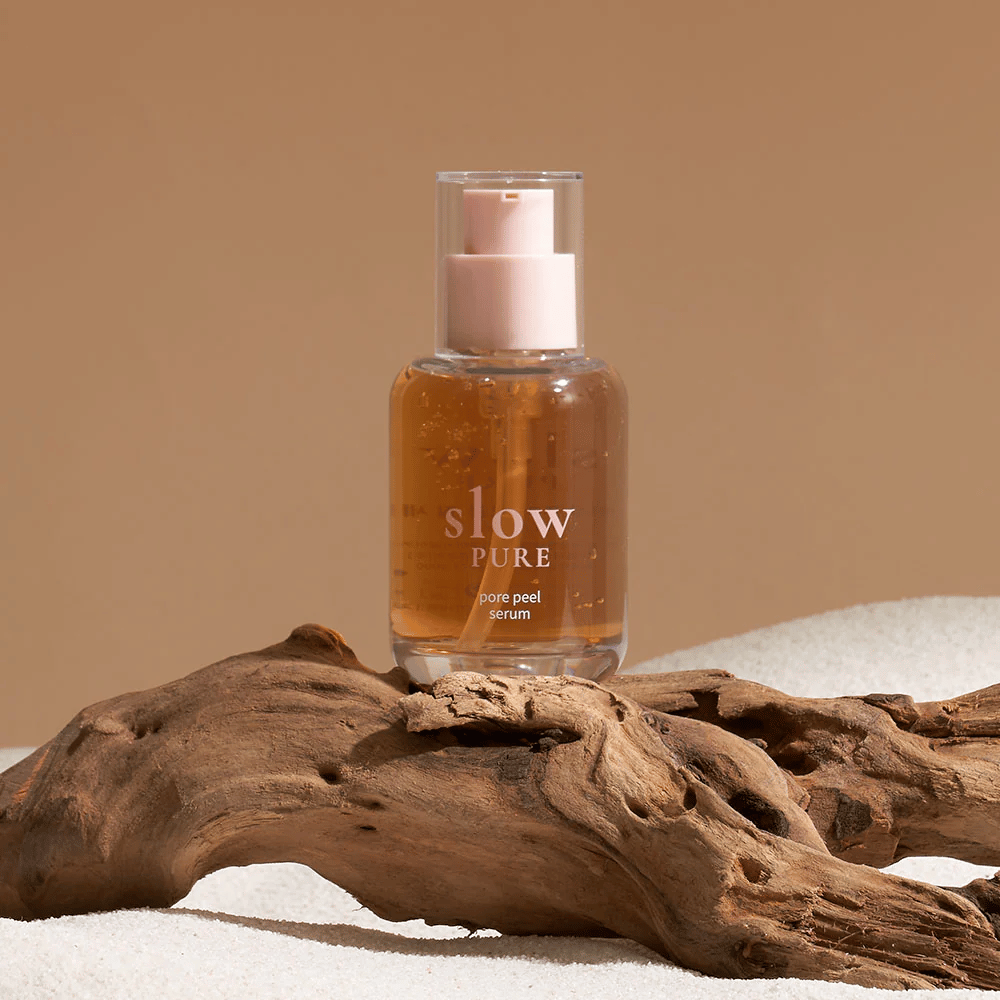Slow Pure Pore Peel Serum placerad på sand med trädetalj i bakgrunden, symbol för naturlig hudvård.