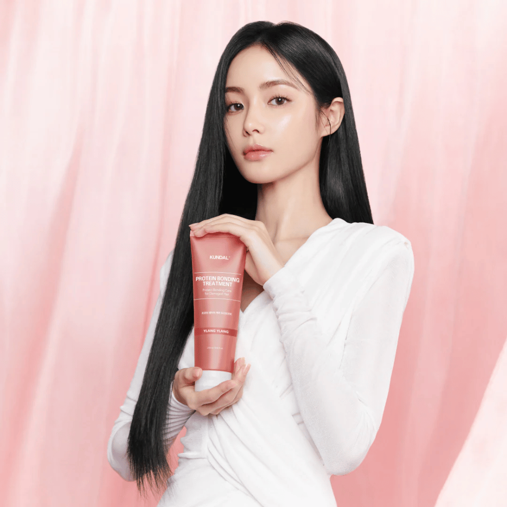 Donna con capelli lunghi e lucenti tiene in mano KUNDAL Protein Bonding Treatment Violet Muguet su sfondo rosa.