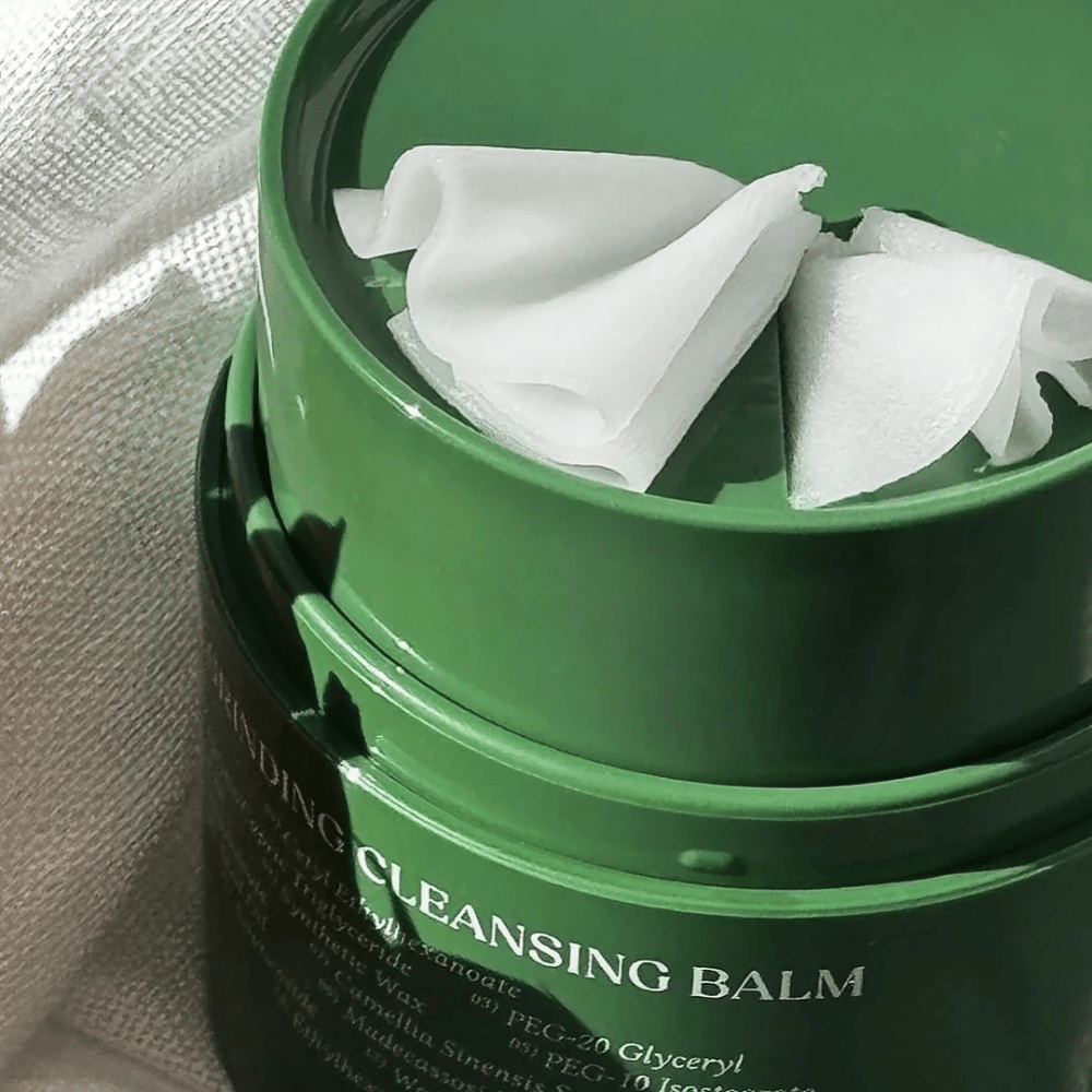 L'immagine mostra un barattolo verde con "Cleansing Balm", che è aperto per rivelare la spatola inclusa e il contenuto morbido. La consistenza cremosa riflette la luce, conferendo una sensazione di lusso ed efficacia nella pulizia.