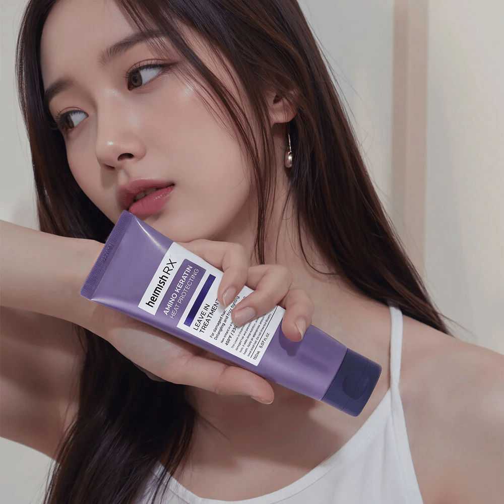 Donna con capelli lunghi e lucenti tiene in mano Heimish RX Amino Keratin Leave-In Treatment – mostra il prodotto come un passaggio curativo e protettivo dal calore nella routine di cura dei capelli.