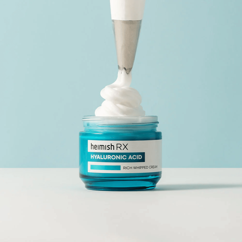 Heimish RX Hyaluronic Acid Rich Whipped Cream in un barattolo di vetro blu, dove un sac à poche applica la crema soffice. Una crema viso idratante con acido ialuronico per un'idratazione intensa e una pelle morbida e piena.